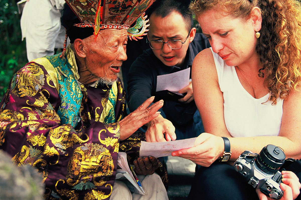 Three people examine a sheet of paper, on dressed in traditional Chinese robe with gold embroidery and headdress, and the other two in Western dress. One holds a film camera.