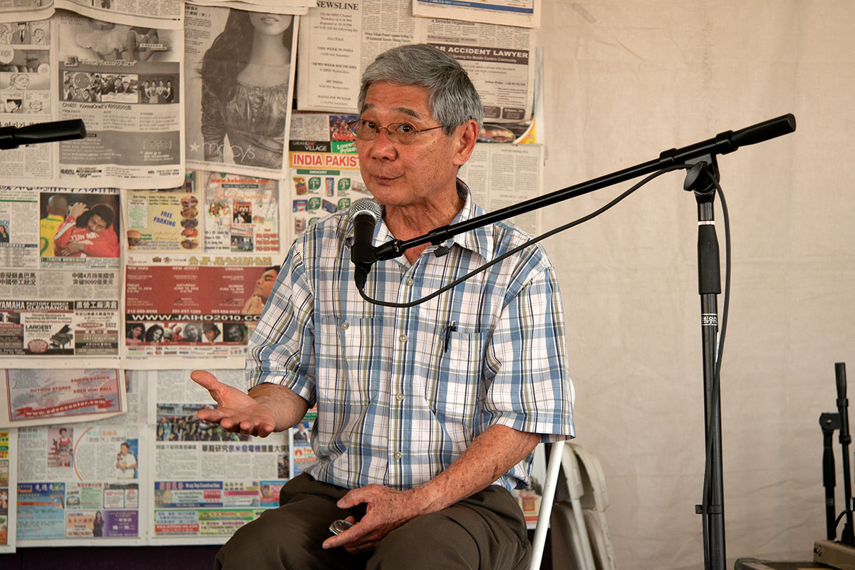 A man sits on stage in front of a microphone, gesturing with one hand. Behind him, a backdrop of newspaper clippings.