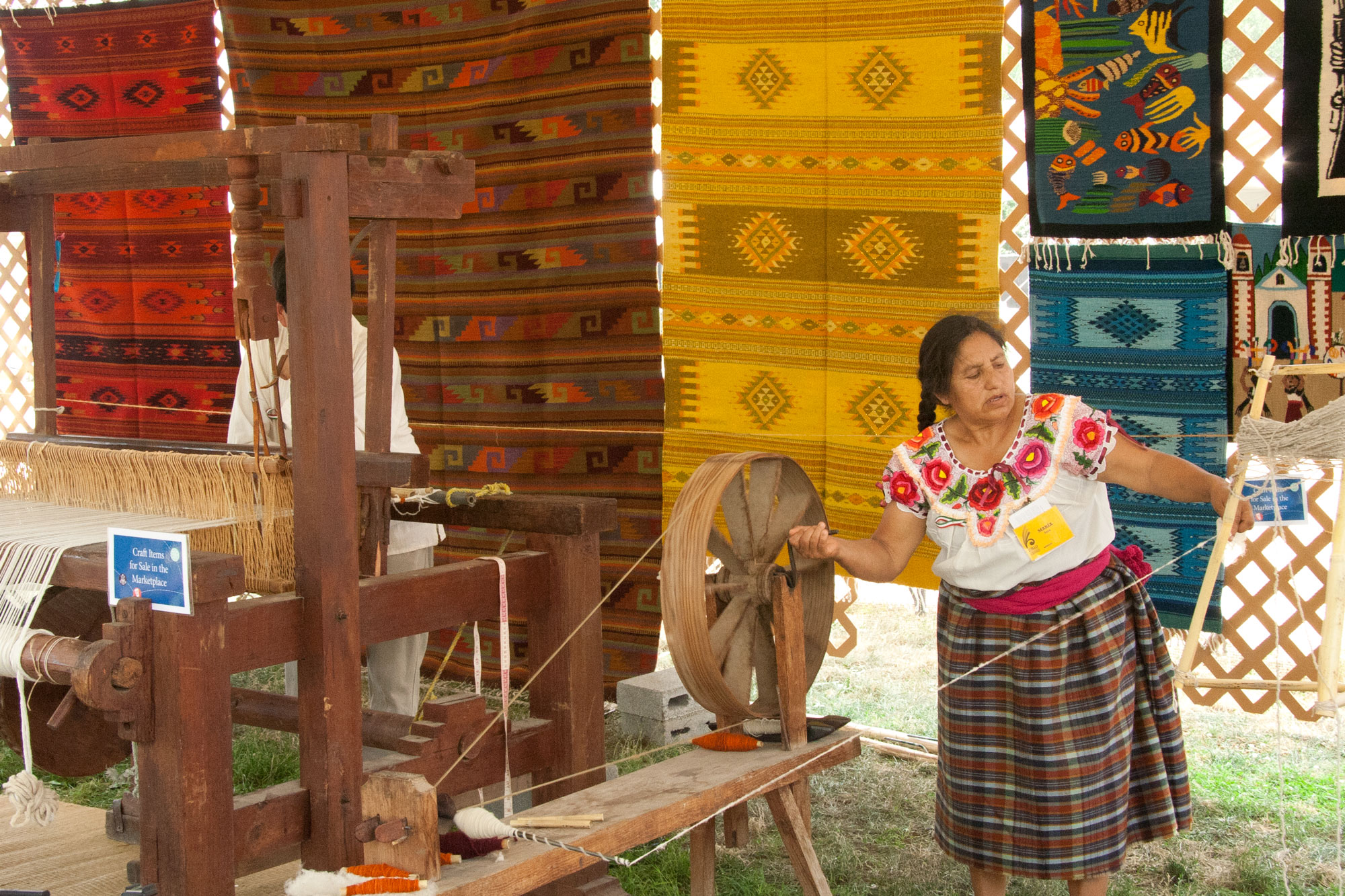 An older woman with dark hair in braids, wearing a traditional Mexican embroidered shirt and plaid skirt, holds wool she is spinning at a spinning wheel. Behind her are brightly colored, woven rugs she and her family have made.