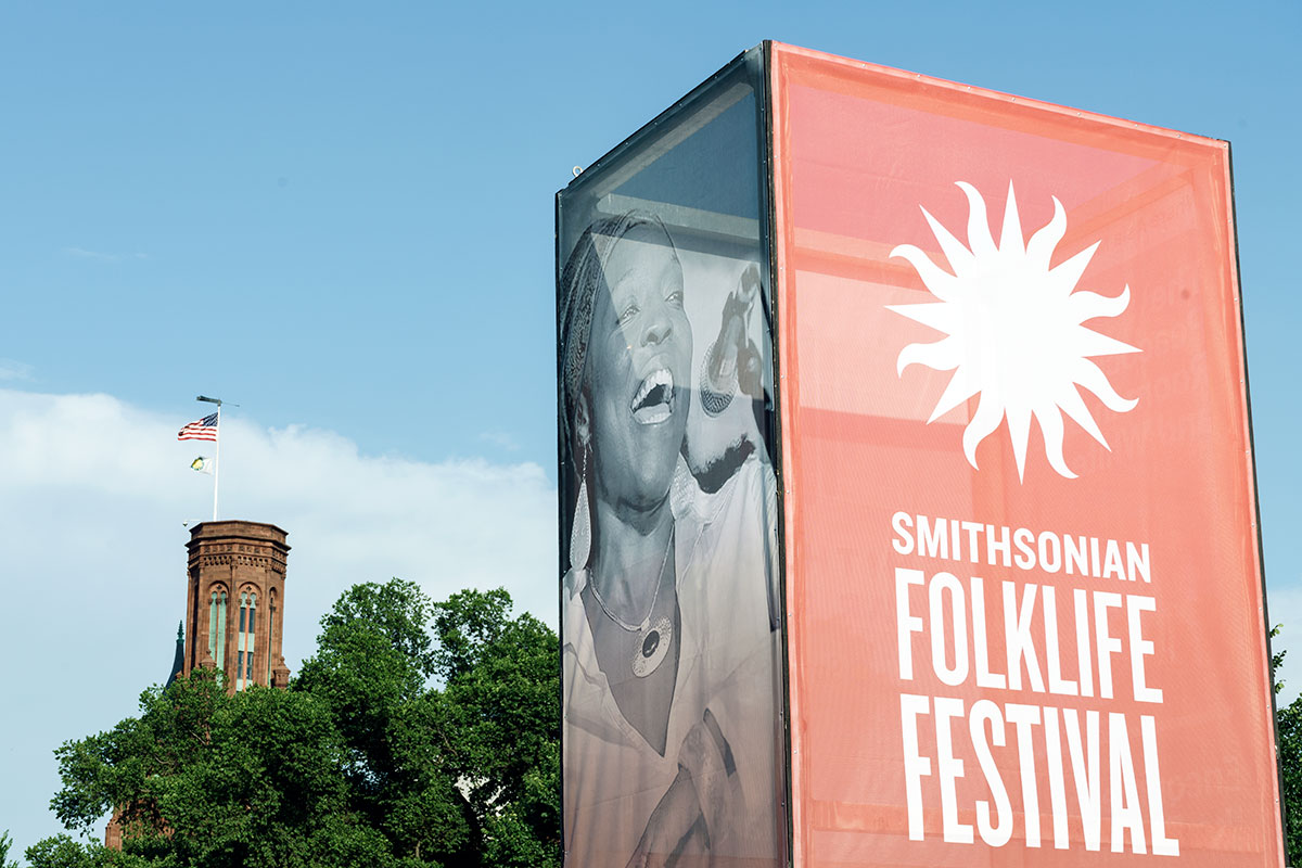 Tall event sign with the Smithsonian Folklife Festival logo in red on one side, photo of a woman singing on another, and the Smithsonian Castle seen in the background.