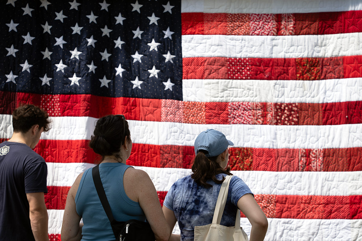 Three people stand in front of a quilt designed like the American flag, with the words of the Pledge of Allegiance stitched into the white stripes and stars in the red stripes. 