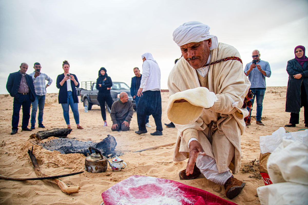 A man dressed in a traditional, white Tunisian turban and garb bends over the sand as he spins bread dough between his hands. A group of onlookers surrounds him in the desert.