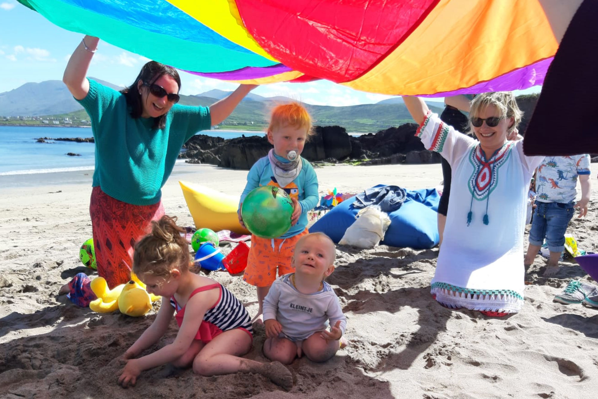 Two women holding a blanket on the beach with two infants playing underneath/Beirt bhan ag imirt le naíonáin.