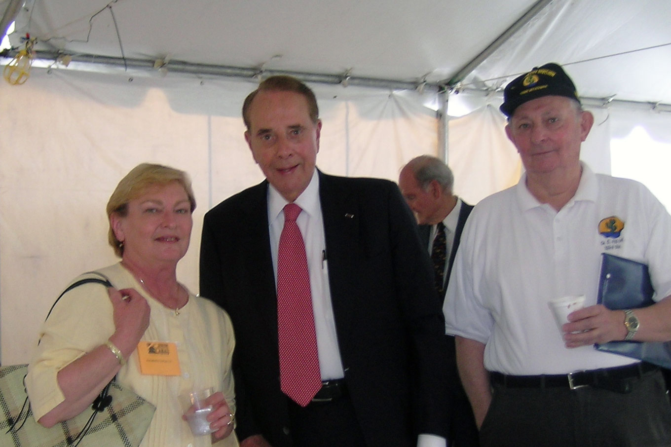 Three people pose under a large white tent. In the center is Bob Dole, dressed in dark suit and red tie, smiling.