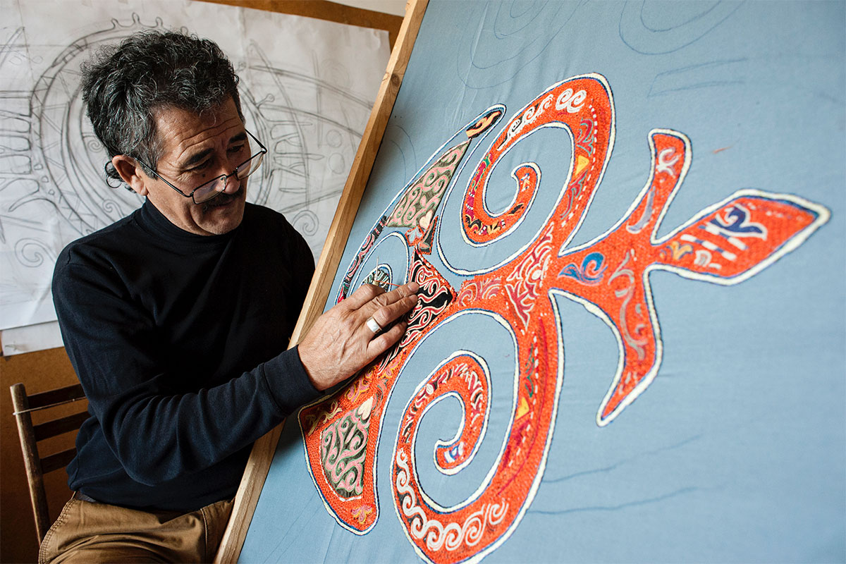 A man inspects and touches a tapestry work in progress: a symmetric, abstract embroidery in orange with multicolored embellishments on a light blue fabric. 