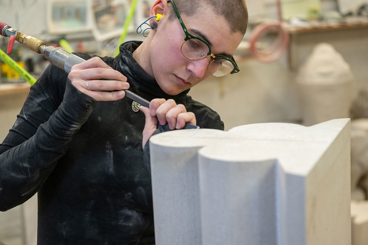 A young woman wearing glasses and earplugs uses a pneumatic chisel on a decorative piece of white stone.