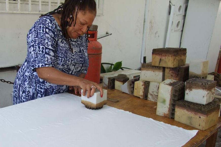 A Black woman stands over a work table, applying a wooden block stamp to a large piece of white paper. Other block stamps are stacked at the edge of the table.