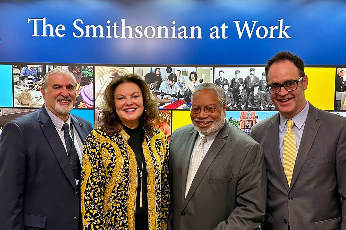 A woman and three men pose underneath a museum display sign that reads, The Smithsonian at Work. The men are dressed in gray suits, and the woman wears a black and gold tunic and gold jewelry.