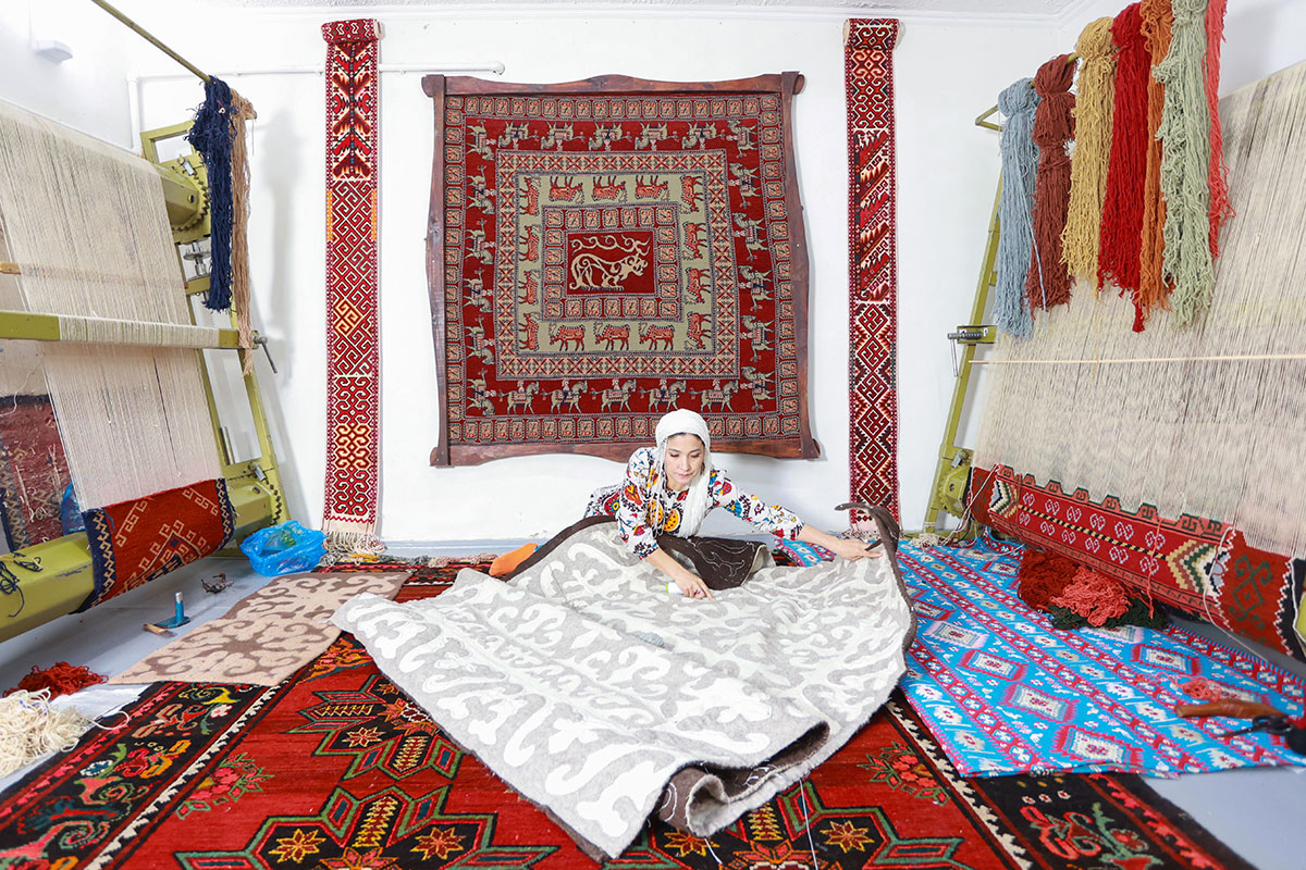 A woman sits on the ground, unrolling a woven gray and white rug. More rugs sit under her and hang behind her, and on either side are two large looms with rugs in progress.