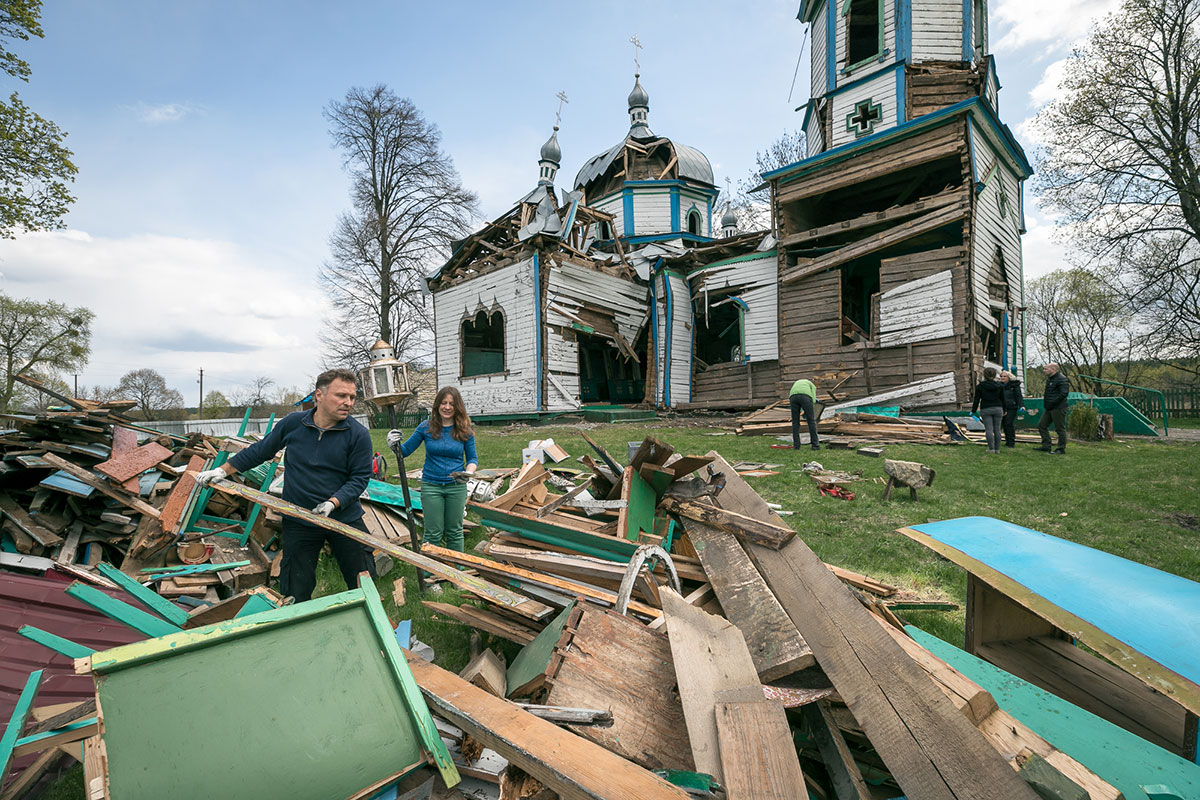In the foreground, two people add planks of wood to a pile of wooden refuse. In the background, a wooden church, painted white with blue trim, that appears to have been bombed.