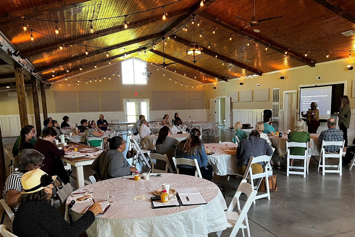 In a large room with wood-paneled ceiling lined with string lights, people sit around white tables as two people in front of a screen give a presentation.