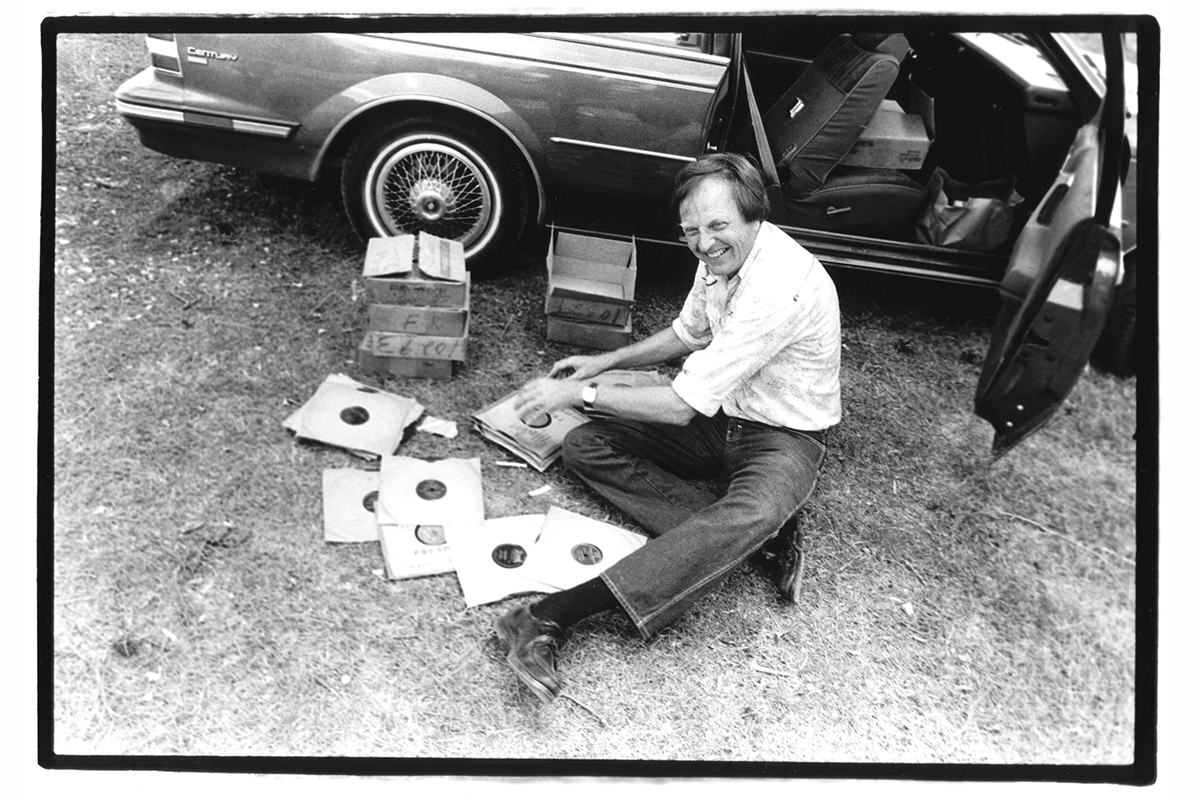 A man sits on the ground next to the open door of a car, sorting a pile a vinyl records and laughing.
