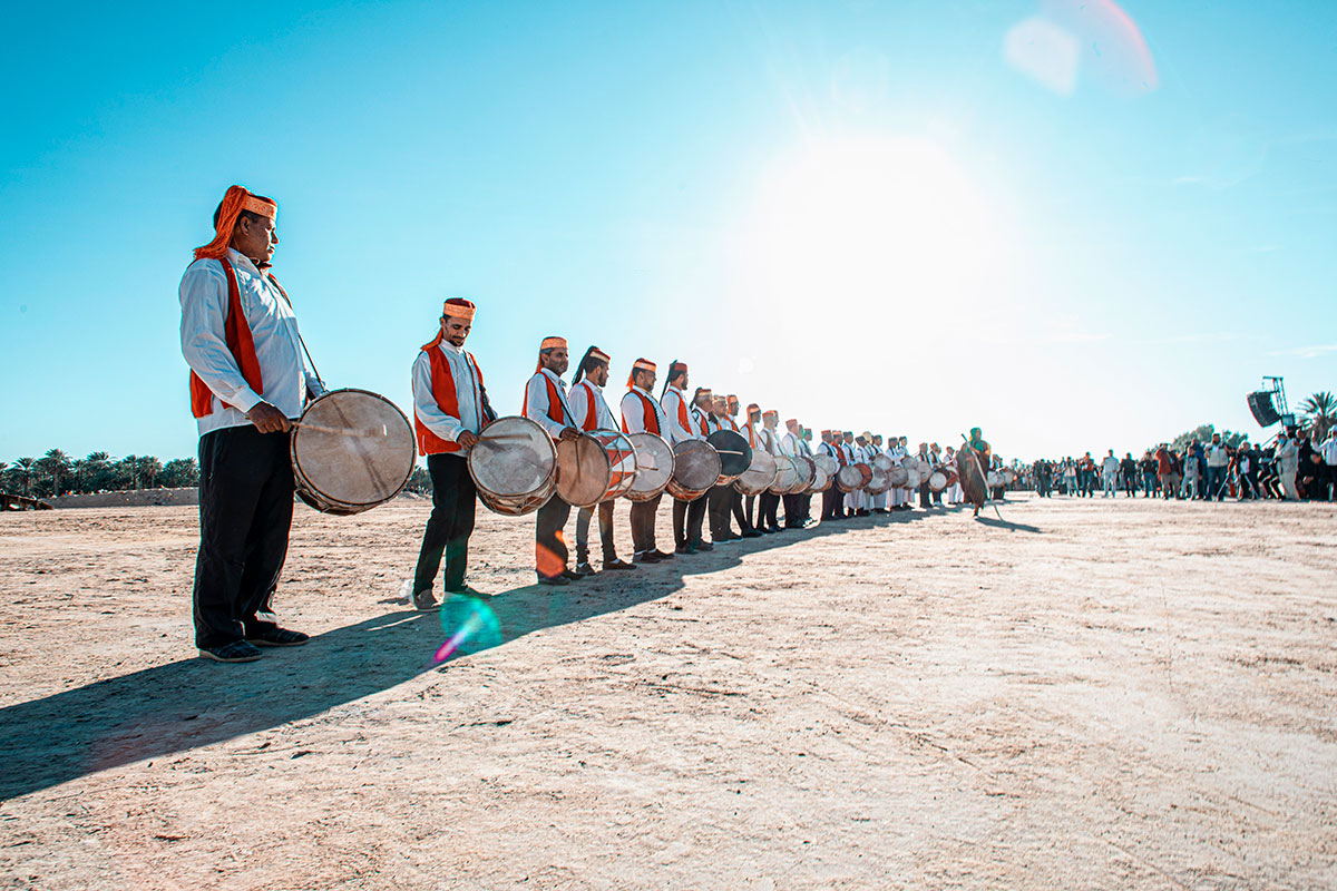 A line of drummers wearing red vests and white shirts stand in the dessert on a clear, blue-skied day.