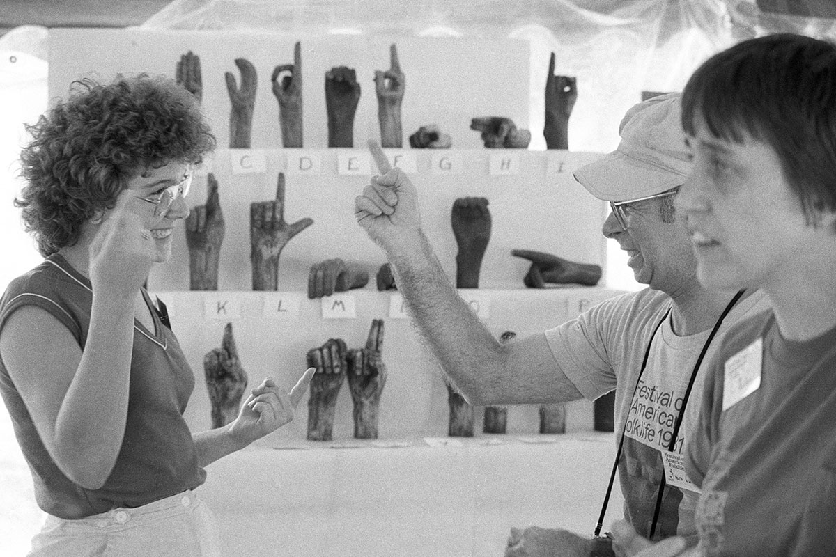A man and a woman communicate using ASL, with a third person in the foreground looking on. Behind them is a display of sculptures of hands, depicting the ASL alphabet. Black-and-white photo.