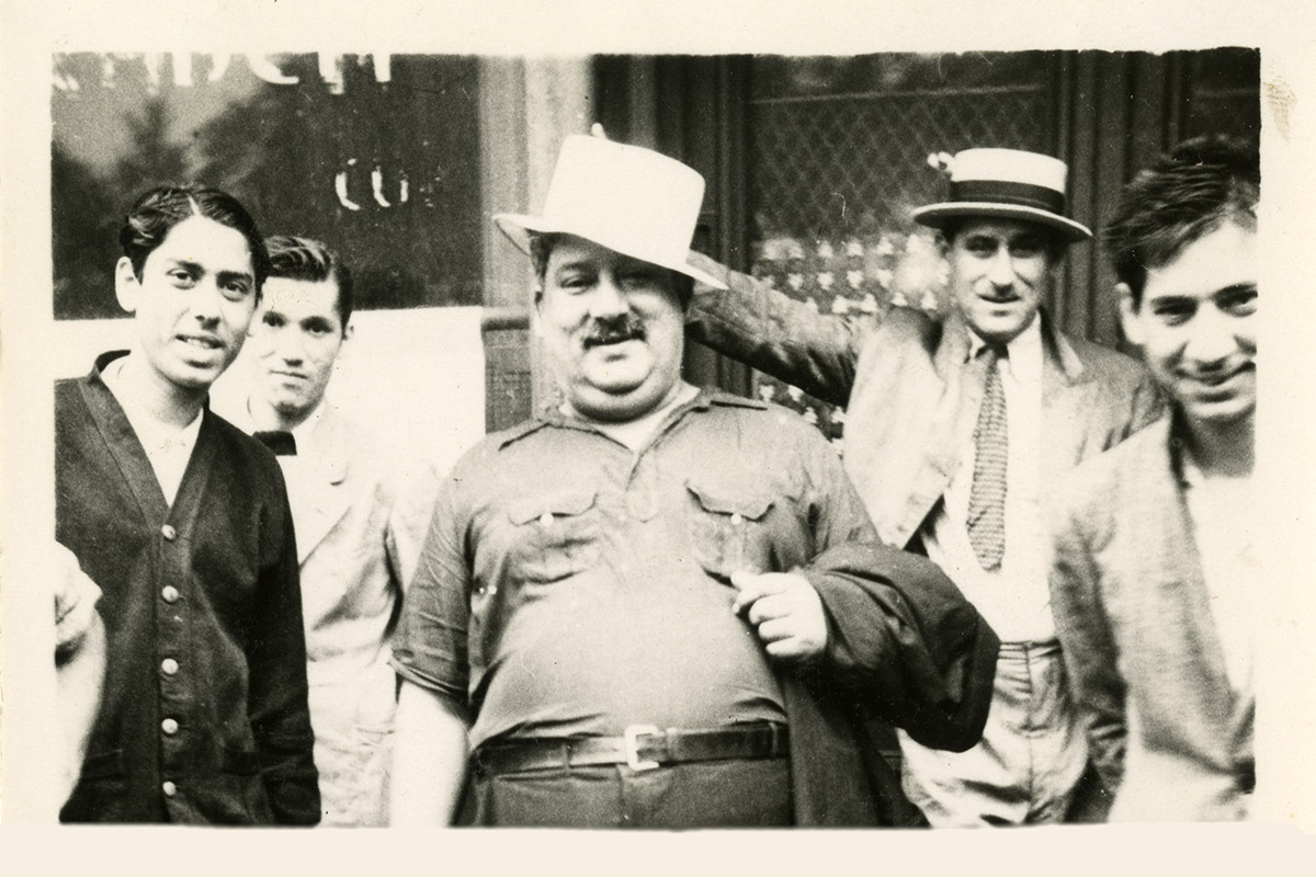 At center, a man in a double-breast-pocket T-shirt and white top hat, with a moustache and holding a jacket over one arm, smiles for the camera, surrounded be four other men in dress sweaters and jackets. Black-and-white photo.