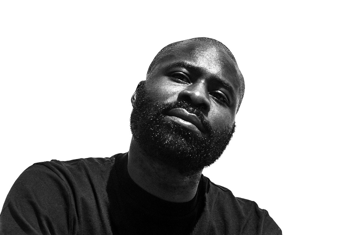 High-contrast, black-and-white portrait of a Black man with a beard and black T-shirt against a pure white backdrop.