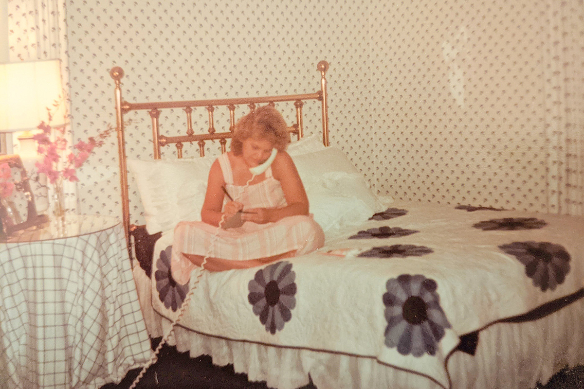 A young woman in a striped sundress sits on a bed, talking on a white telephone. The bedspread is a white quilt with blue flowers.