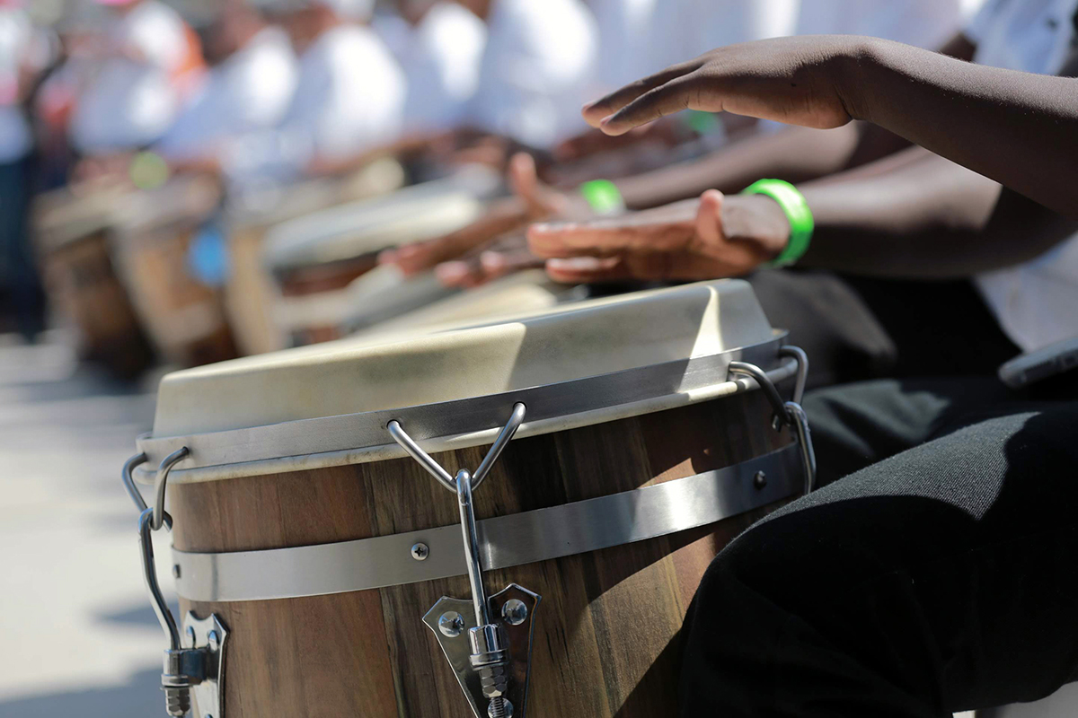 Close-up on a pair of Black hands playing a drums. In the blurry background, a whole line of seated drummers.