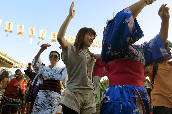 People in kimonos and plain clothes dance in a line together, arms outstretched above their heads. Paper lanterns hang in the background.