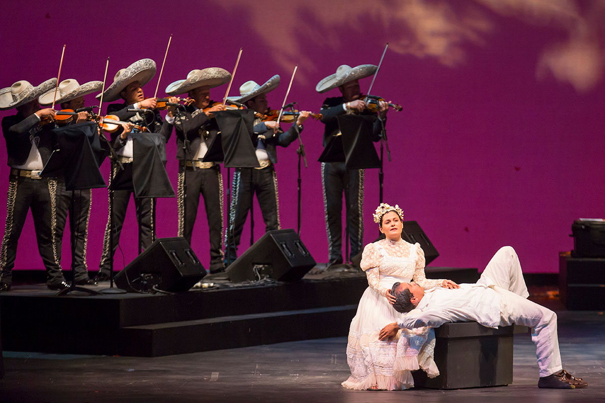 On a stage with a purple background, a man dressed in white lies on  his back while a woman, also in white, puts her hands to his head and chest. Behind them, six members of a mariachi band in matching dark suits and large white sombreros, all playing violin.