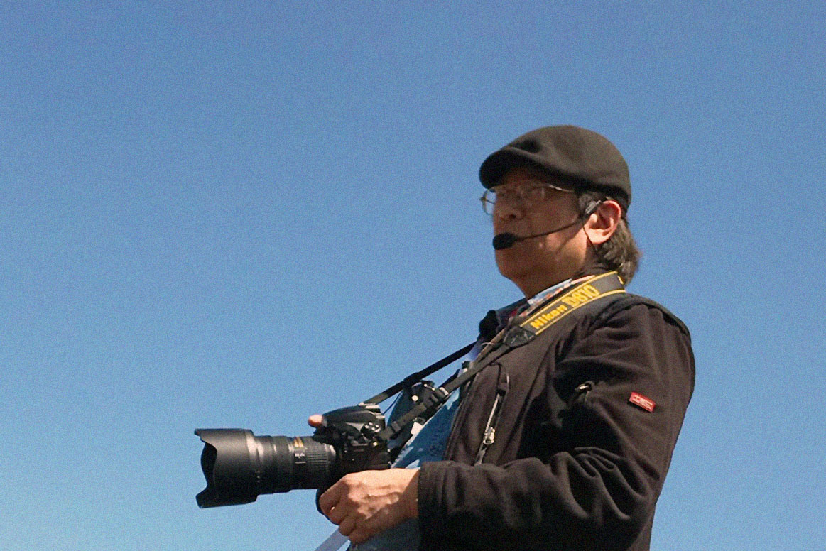 An older Asian man standing against blue sky background, holding a DSLR camera with big lens and a headset.