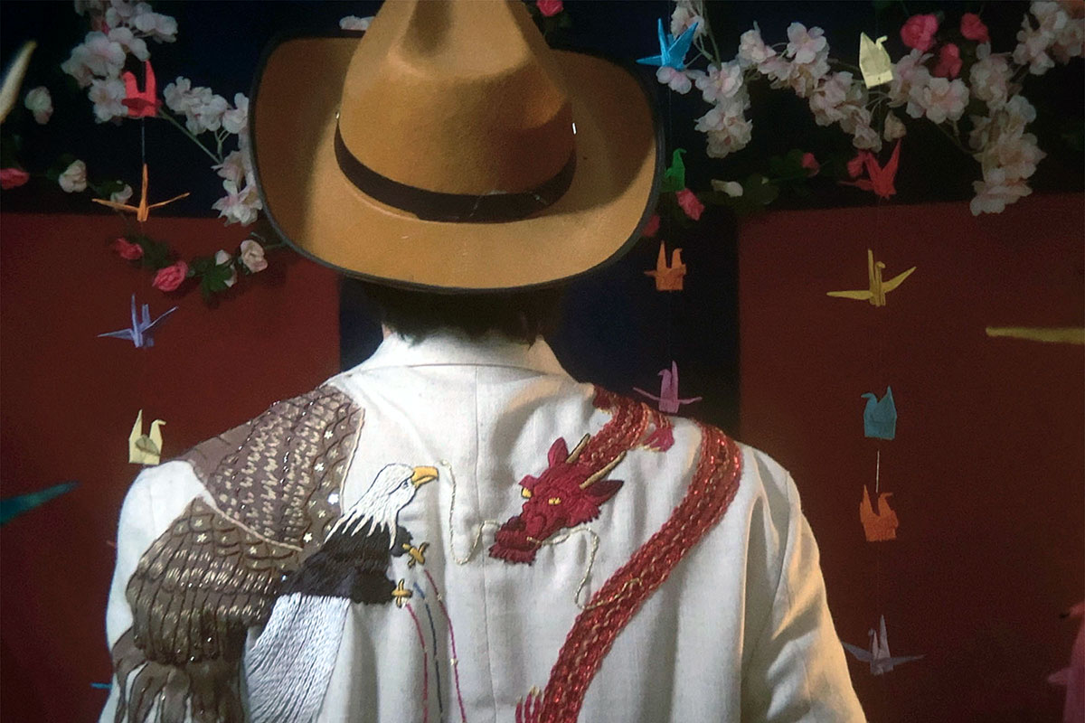 The back of a person wearing a brown fedora and white jacket embroidered with a bald eagle on the left shoulder fighting a red dragon on the right. Origami cranes are hanging in the background.