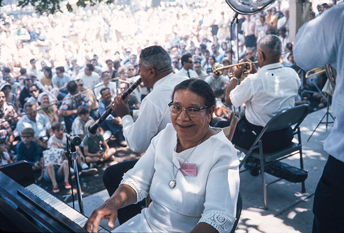 A jazz band plays on an outdoor stage for a huge crowd. The woman pianist in the foreground smiles for the camera.