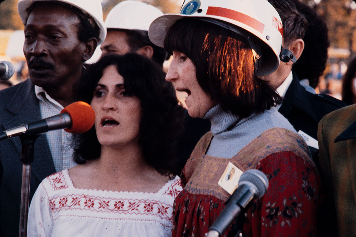 A group of people, with two women in the forefront, sing together outdoors. Some of them, including one of the women, are wearing hard hats.