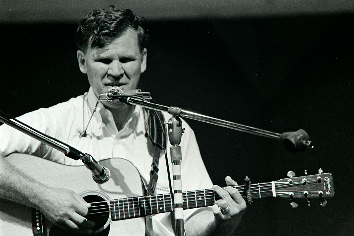 A white man plays acoustic guitar on stage, with two microphones and a harmonica in front of him and a harmonica holster around his neck. Black-and-white photo.