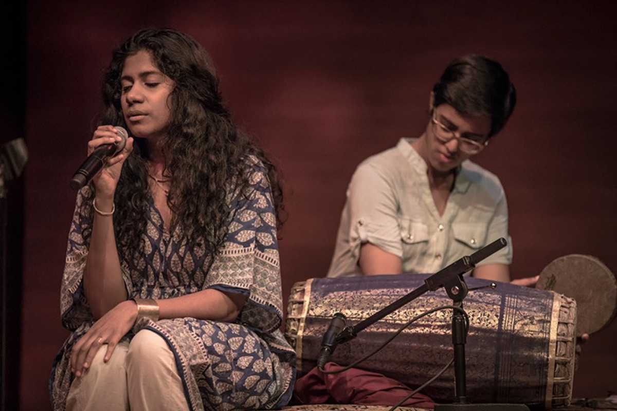 On the left, a woman with long wavy dark hair and patterned blue blouse sings into a microphone, seated, eyes closed. On the right, another person sits behind a long barrel-shaped drum, holding a hand drum, looking down.