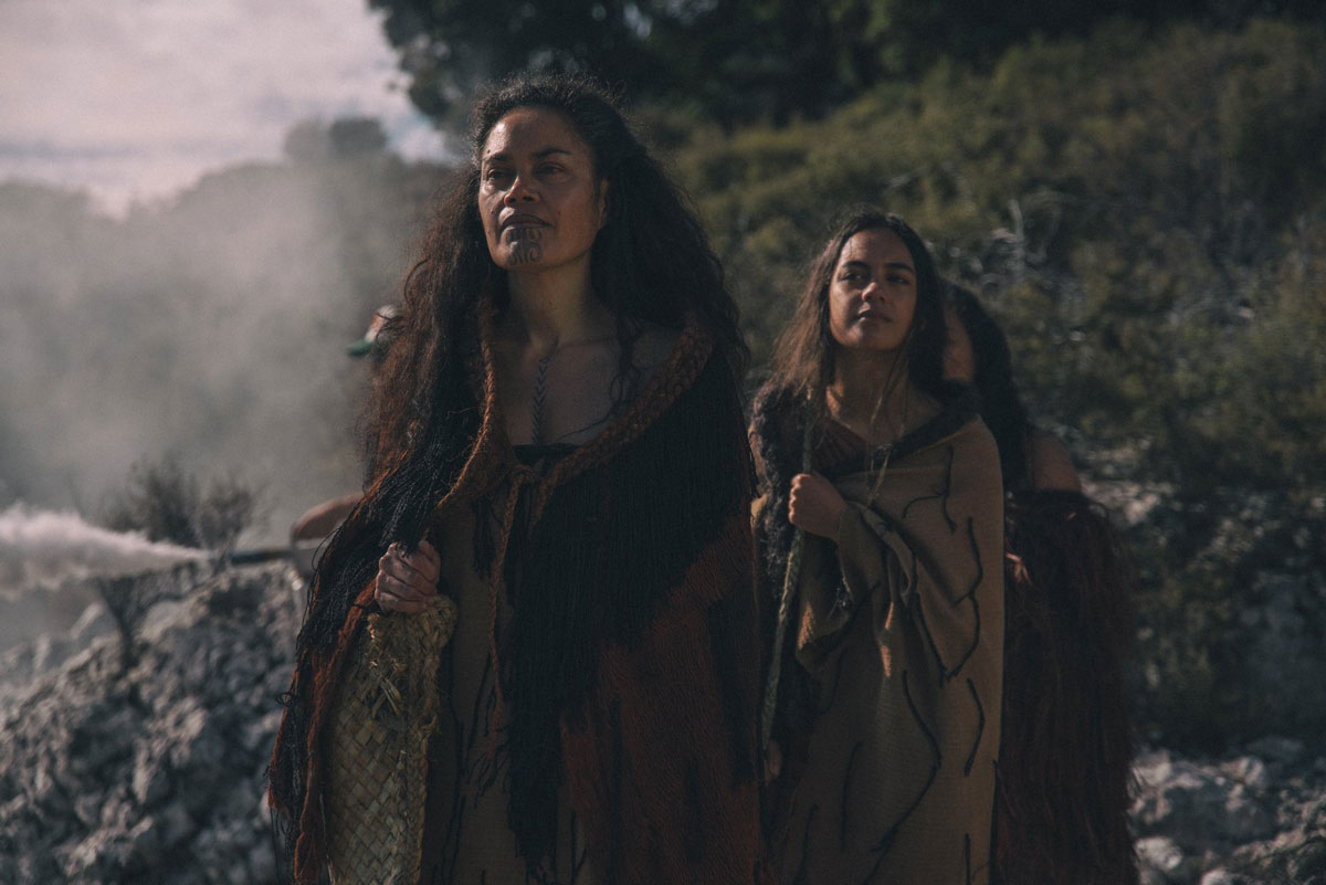 An older woman and her daughter stand one in front of another wearing traditional Maori dress, walking up a mountain.