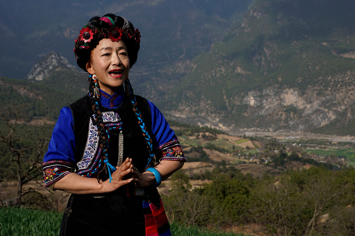 A woman in traditional Naxi dress of blue and black embroidered blouse and floral headwrap sings outside, a mountainous landscape behind her.