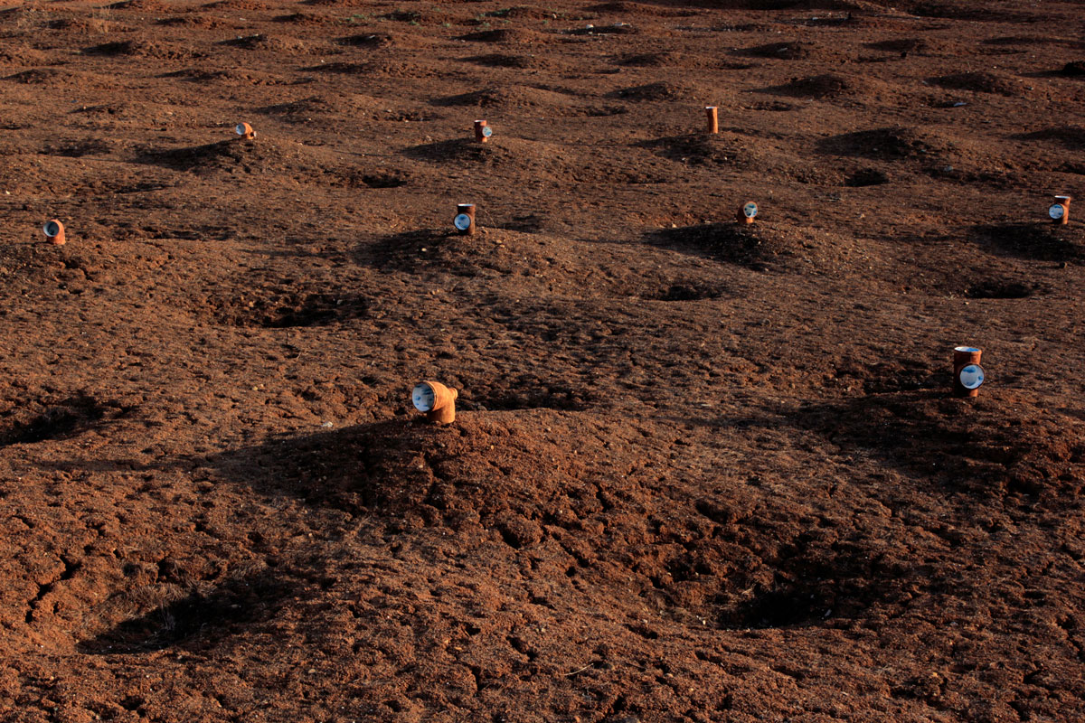From a distance are pictured an arrangement of nine rusty pipes, bent and opening at various angels are placed in the dark soil. The visible openings are painted to appear like a blue sky with white, fluffy clouds.