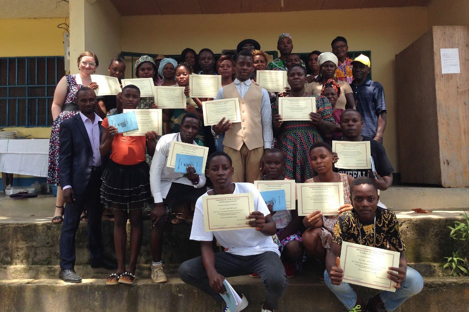 Iyasa youth, their family members, and teachers stand together in front of a yellow school building. The youth hold certificates showing their completion of the 2018 language workshop.