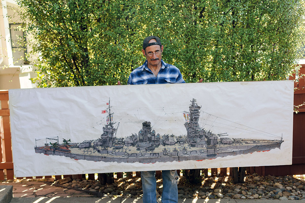 A man holds up a detailed drawing of a gray battleship drawn on a sheet of butcher paper, longer than the man is tall.
