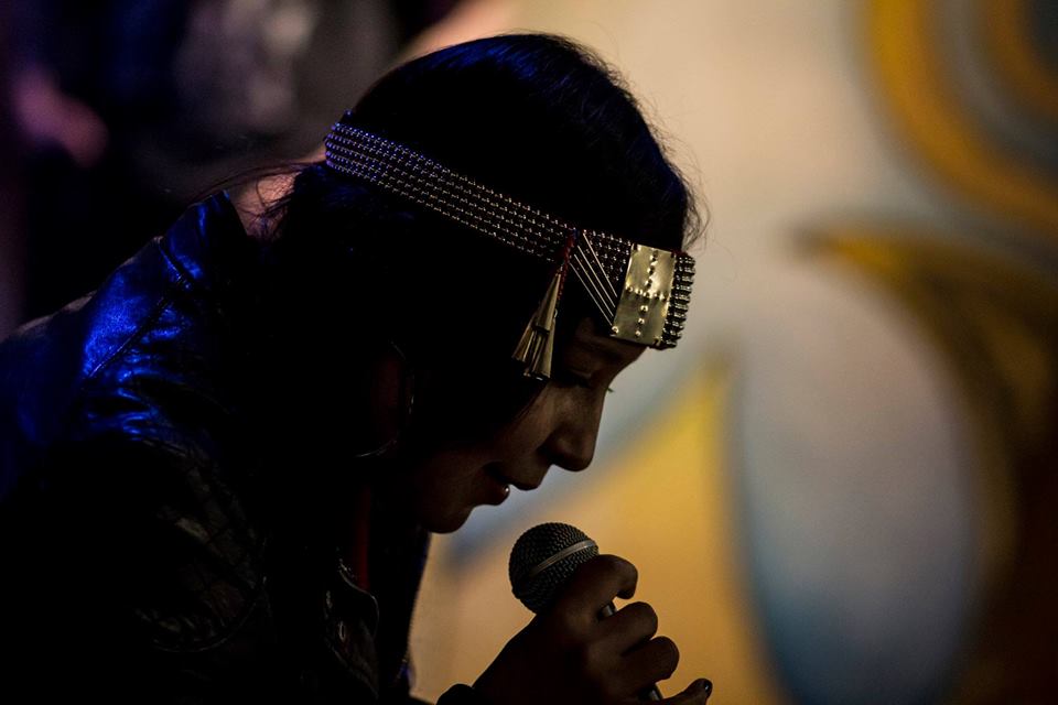 A performer in profile, hunched over and holding a microphone to their mouth, wearing a silver beaded headband.