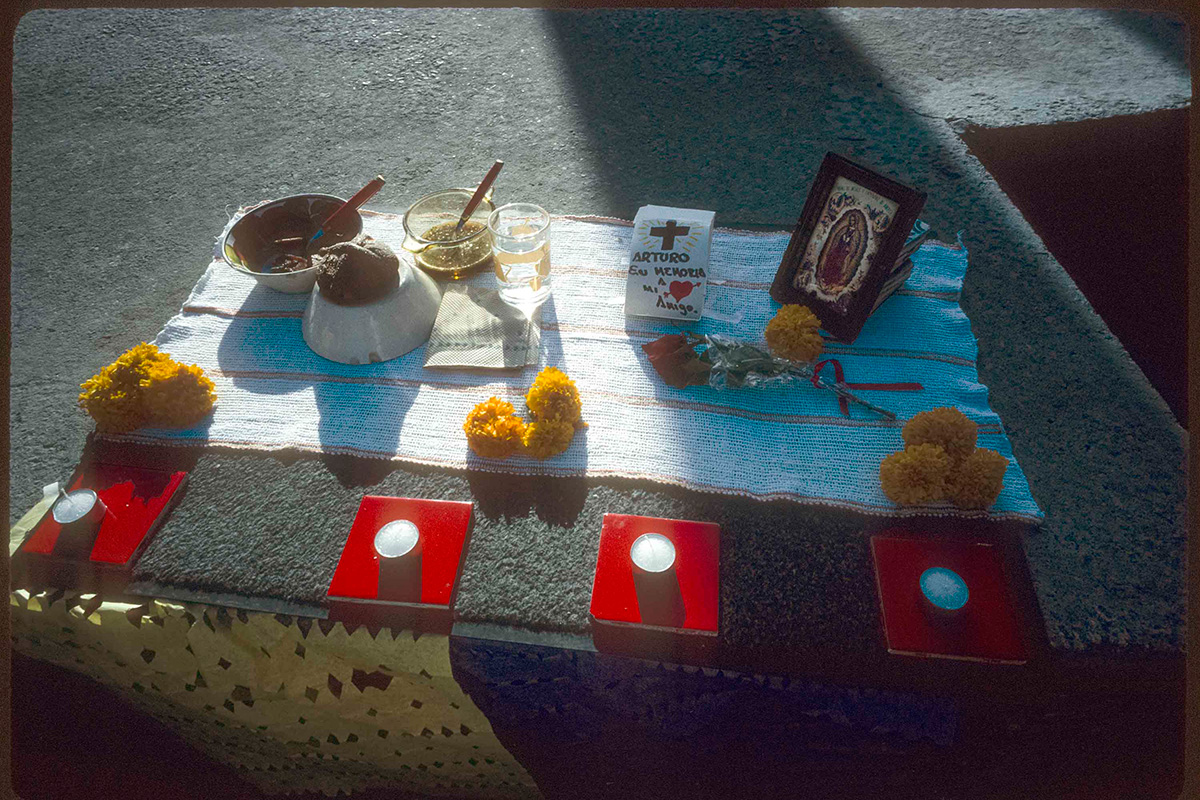 A small offering placed on a cloth on the ground: yellow marigolds, bowls of food, a framed image, and candles.