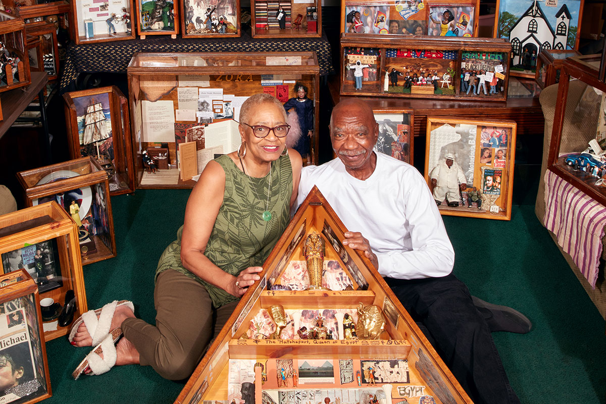 A man and woman pose sitting on the ground, surrounding by wooden diorama boxes.