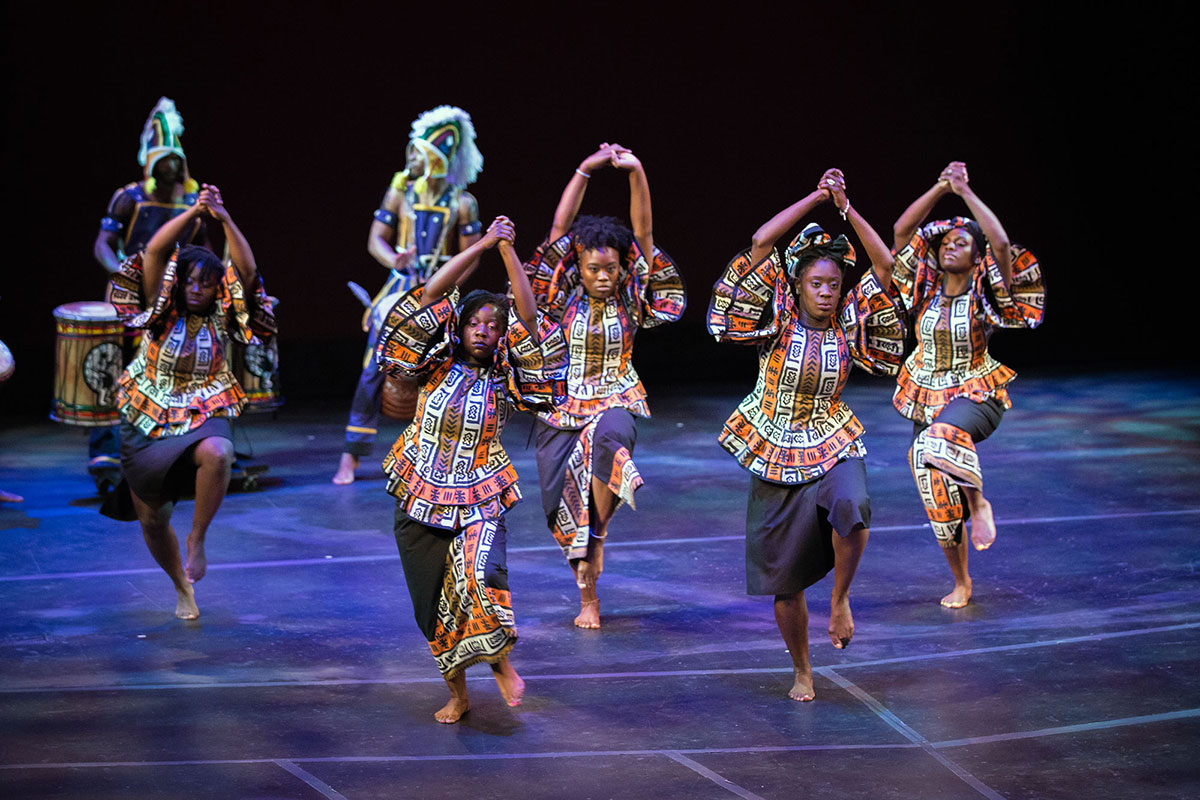 Seven African American people dance on a stage, hands clasped overhead, wearing traditional printed blouses and skirts. Black backdrop.