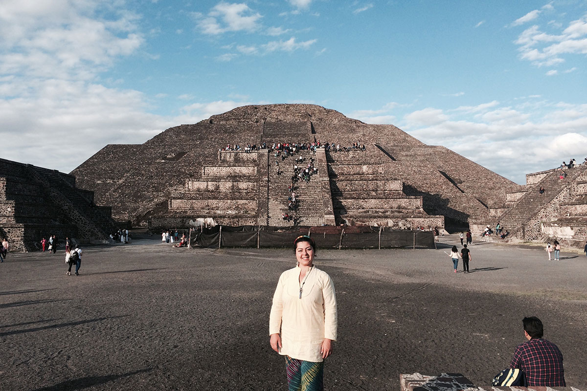 A woman in white blouse and tie-dyed pants poses with a brick pyramid in the background. People climb the steps of the distant pyramid.