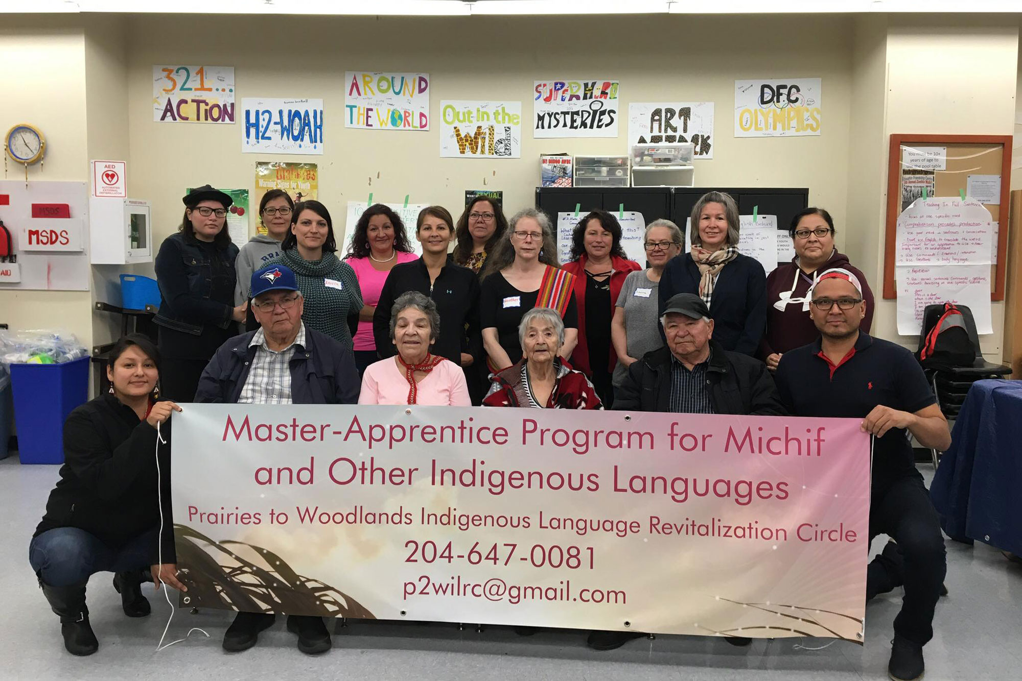 A multigenerational group of seventeen people stand in three rows for a photo. They are indoors, in a classroom and are holding a sign that reads: Master-Apprentice program for Michif and Other Indigenous Laguages.