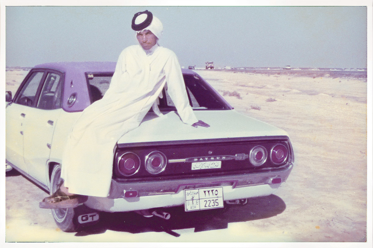 Man in long white robe with black tie securing the white headwrap poses on the trunk of an old white Datsun in a desert landscape. Old film photo with faded colors.
