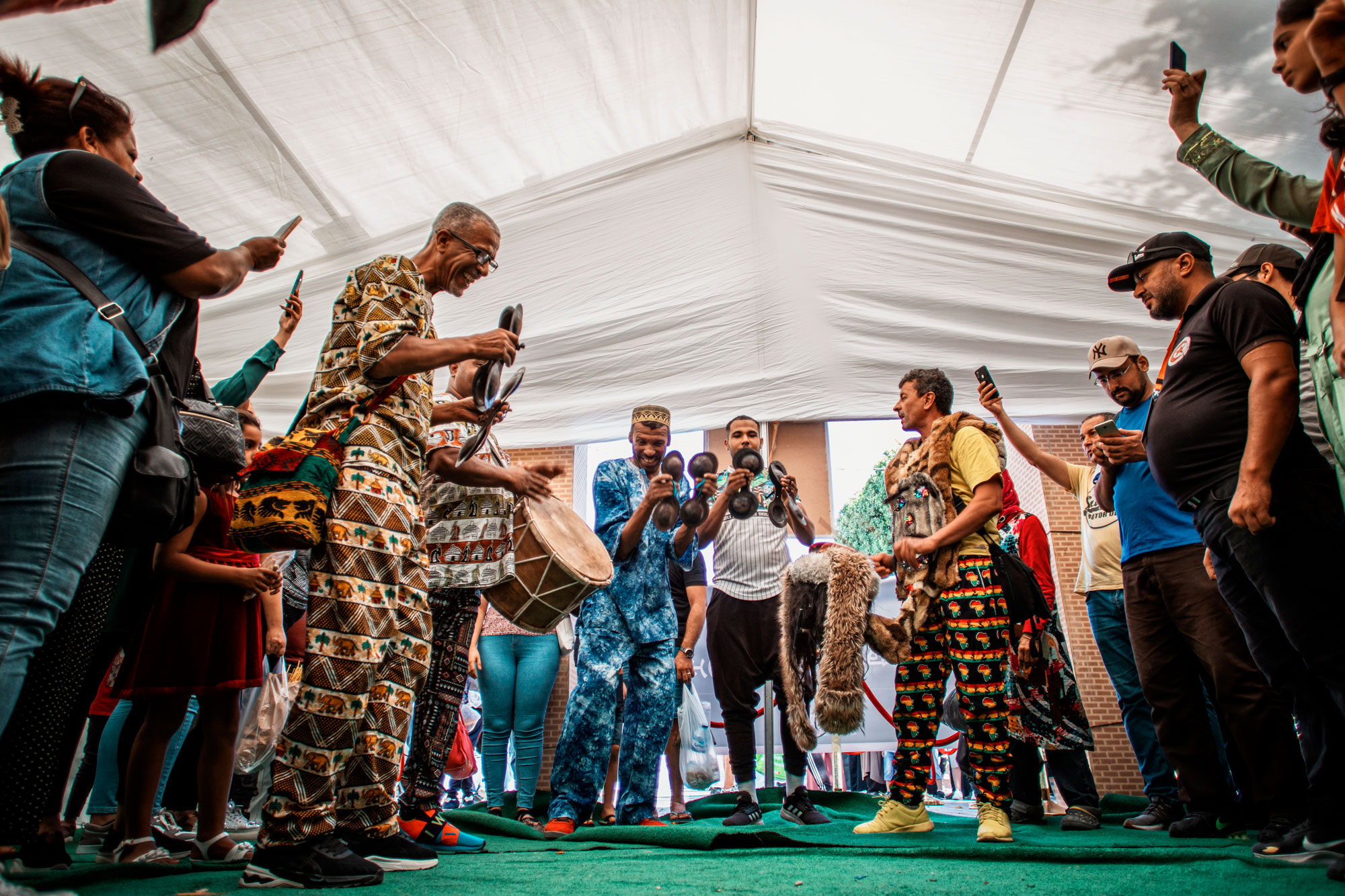 Four men stand together on a tented stage playing percussive instruments. They are dressed in brightly patterned clothes and are filmed by people holding cellphones.