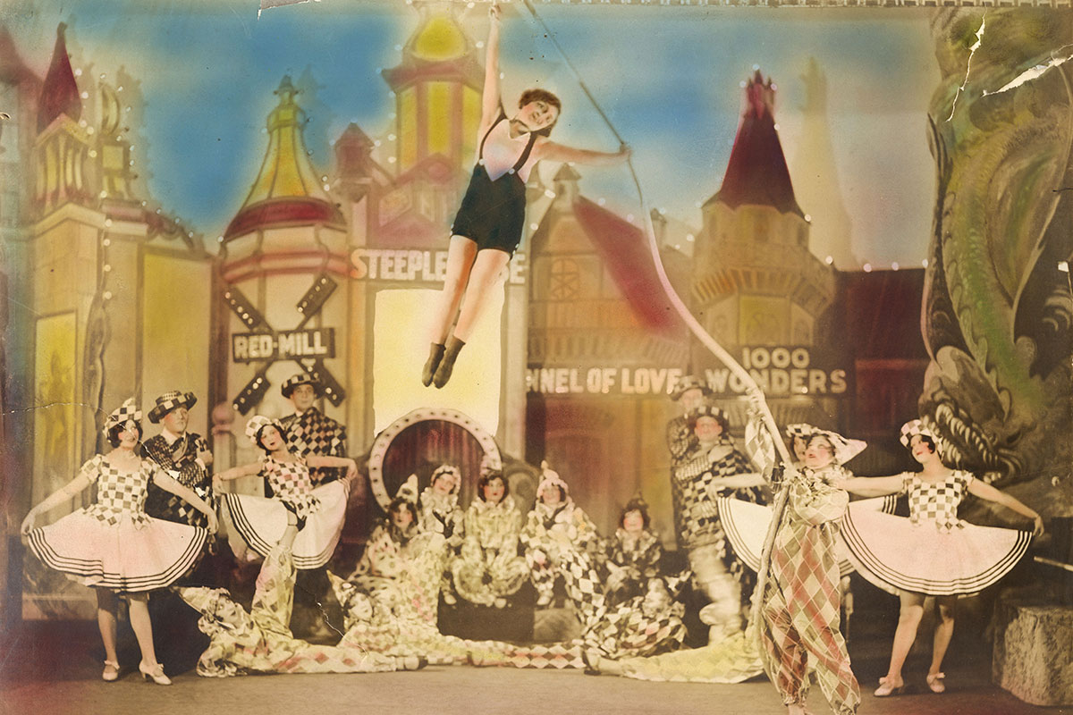 Tinted photograph of a theater production, with lead woman flying on a rope above the rest of the cast, dressed like circus clowns.