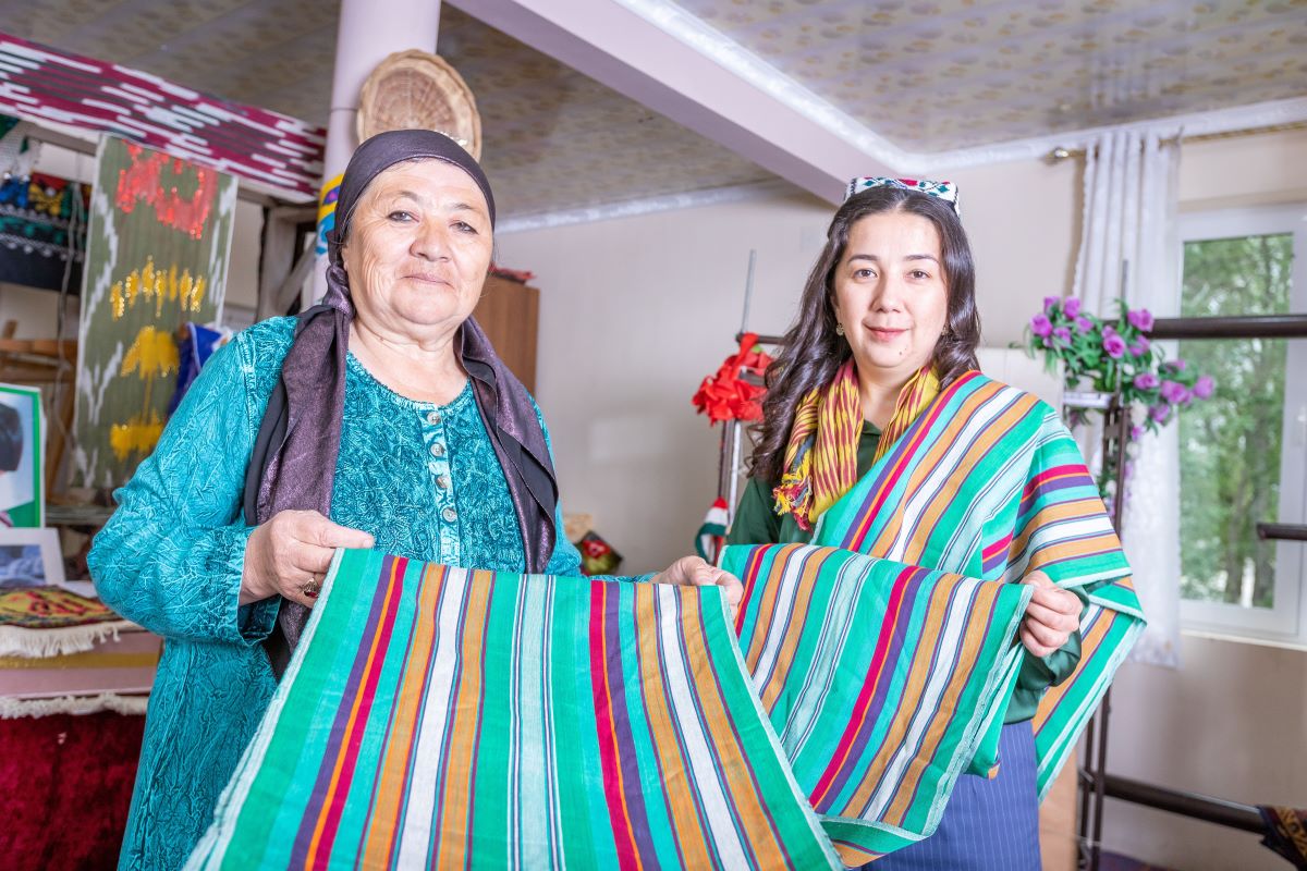 Nigina Ikromi and another artisan smile softly at the camera while holding a colorful piece of cloth.