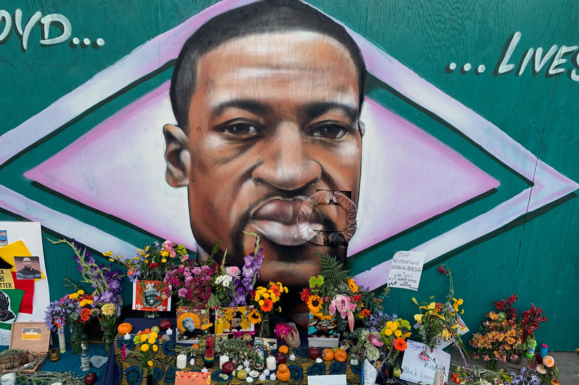 Realistic portrait of a Black man painted on a wall, over a pink diamond design on green background. On the ground in front of the mural, vases of colorful flowers, candles, photos, and written messages.