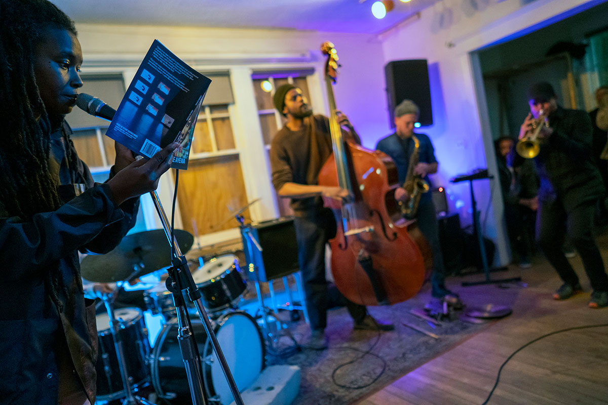 Four performers (vocalist, upright, bassist, saxophonist, and trumpeter) playing in a room of a house. Drummer is out of frame.