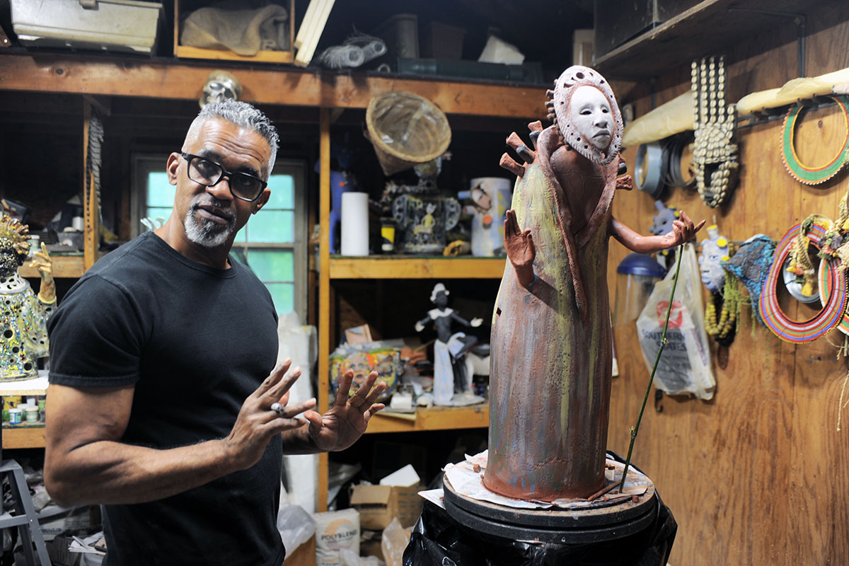 In a wooden studio with shelves and walls full of art materials, a Black man with short graying hair and goatee, black glasses, and black T-shirt gestures toward a sculpture of a standing figure with white mask and outstretched hands.