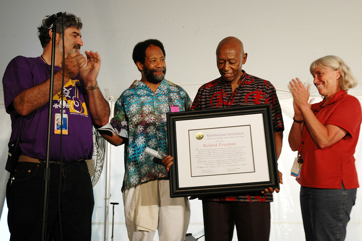 Four people stand on stage; a man holds a framed certificate with a Smithsonian gold seal and his name: Roland Freeman. Two others clap for him.