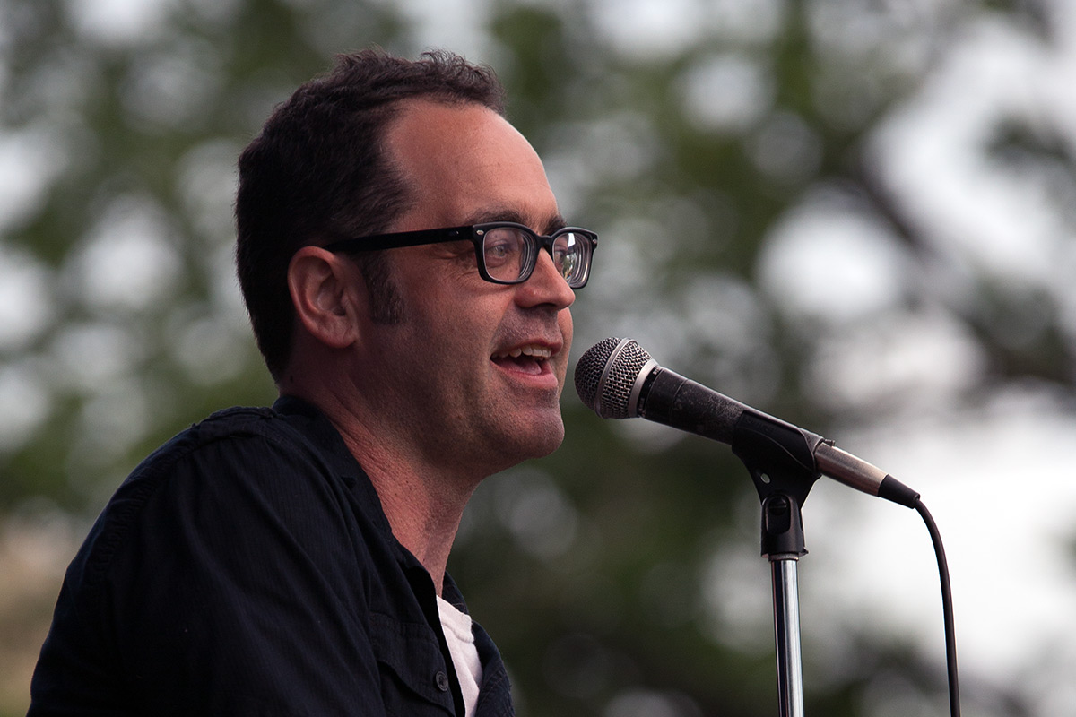 Man with short dark hair, glasses, and black shirt speaks into a microphone on an outdoor stage, trees blurred in the background.